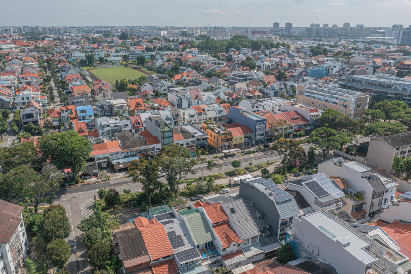 Aerial view of a dense residential area with houses, roads, trees, and a field. High-rise buildings are visible in the distance.